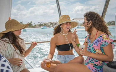 Group enjoying catamaran cruise with drinks, Punta Cana coastline in background.