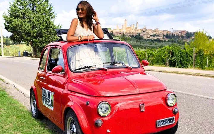 Red vintage Fiat 500 on Chianti road with San Gimignano skyline in background.