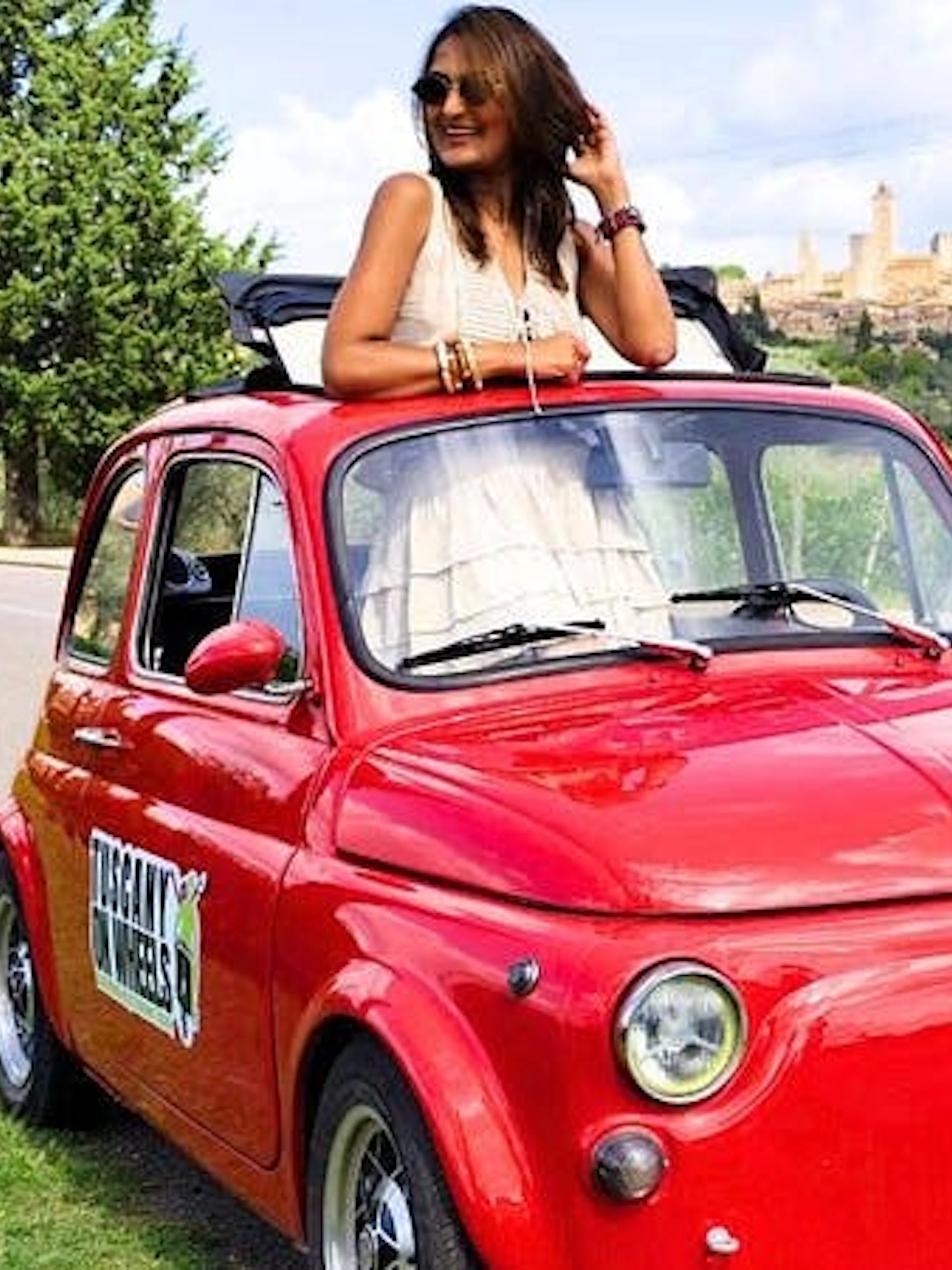 Red vintage Fiat 500 on Chianti road with San Gimignano skyline in background.