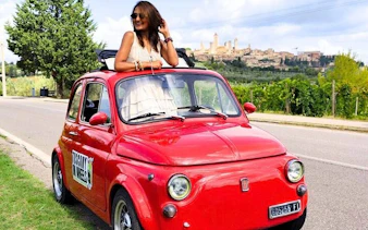 Red vintage Fiat 500 on Chianti road with San Gimignano skyline in background.