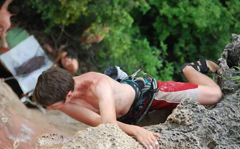 Rock climber ascending limestone cliff at Railay Beach, Krabi, with lush greenery in the background.