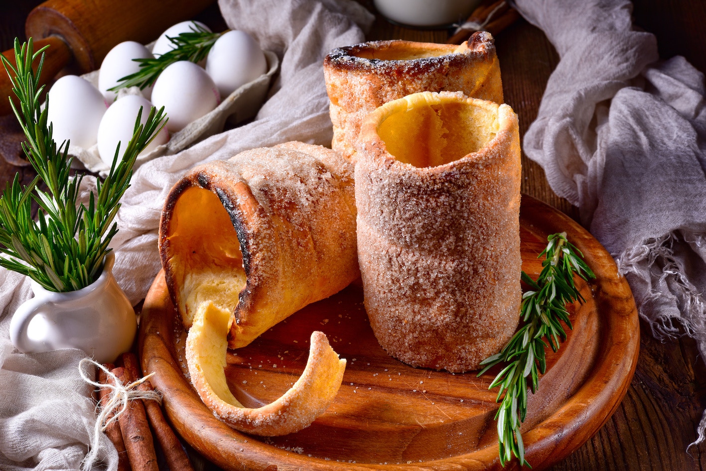 Trdelník pastries on a wooden plate with rosemary and eggs in the background.