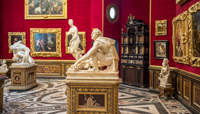 Interior view of Uffizi Gallery showcasing Renaissance sculptures and paintings, with detailed marble flooring and red walls adorned with golden framed artworks, highlighting the cultural richness of Florence