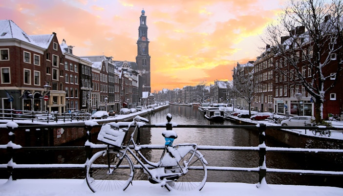 Snow-covered bicycle on an Amsterdam canal bridge at sunset, with Westerkerk in the background.