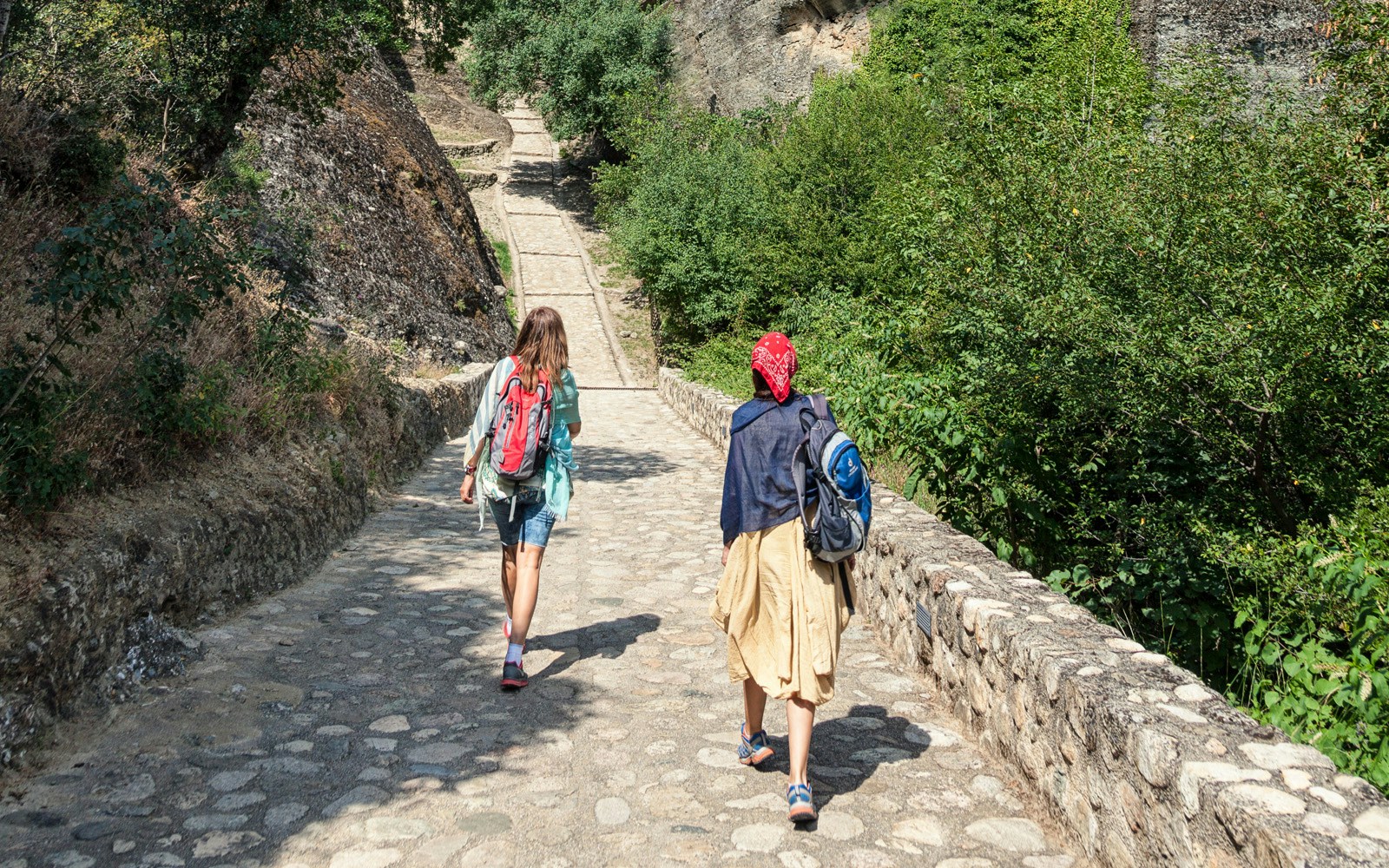 Hikers walking on a stone path surrounded by greenery in Meteora, Greece.