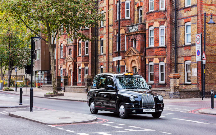 Black taxi driving on a London street with historic buildings in the background.
