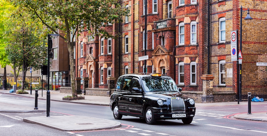 Black taxi driving on a London street with historic buildings in the background.
