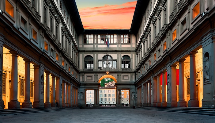Dawn breaks over the Uffizi Gallery, casting a warm glow on the historic columns and sculptures, with the Florentine skyline peering through the archway