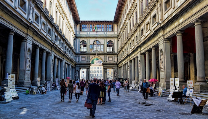 Art lovers and tourists stroll through the bustling courtyard of the Uffizi Gallery, flanked by local artists' displays under Florence's open sky