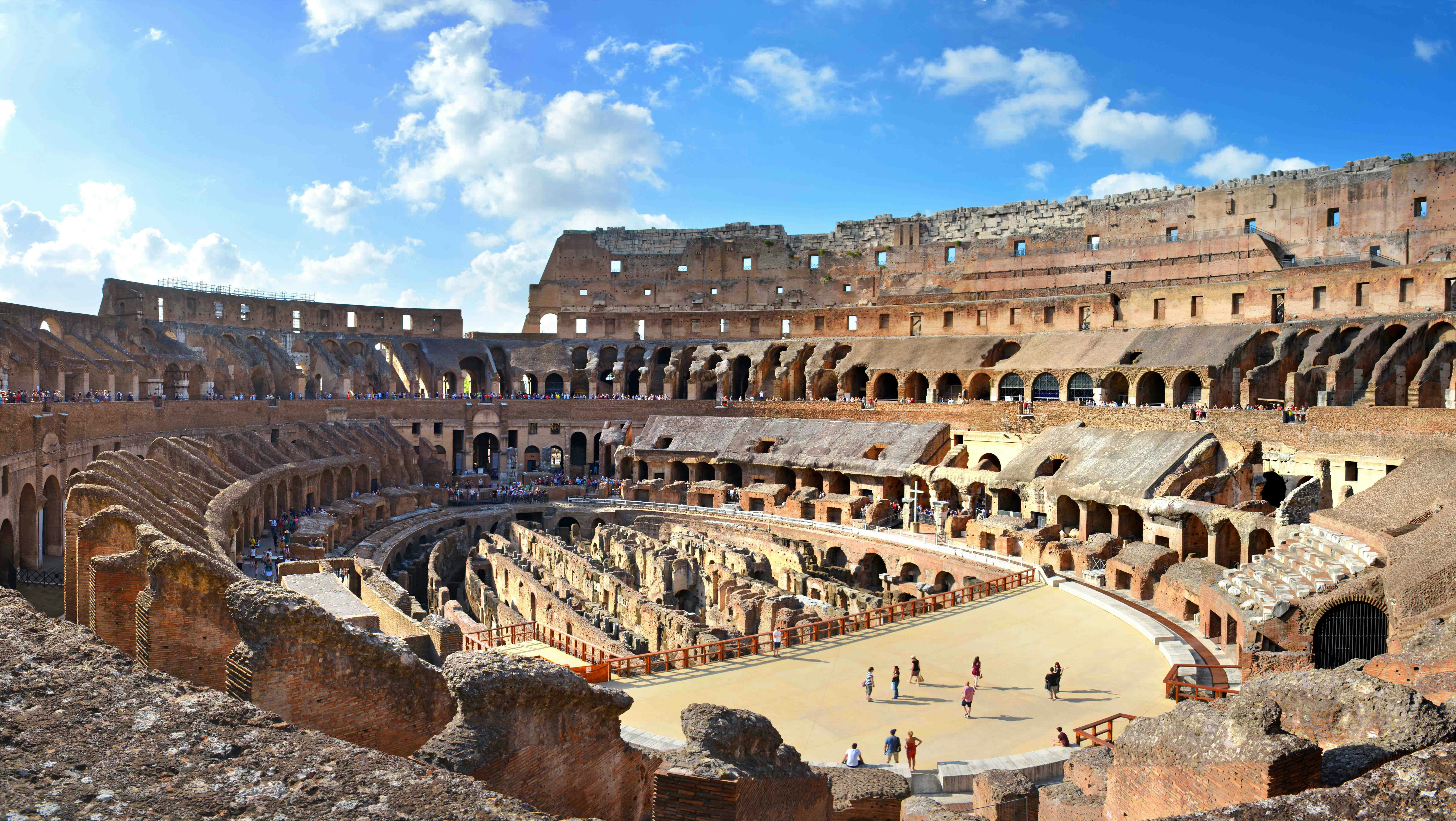 Panoramablick auf das Innere des Kolosseums in Rom mit Besuchern, Ruinen und der rekonstruierten Arena unter blauem Himmel.