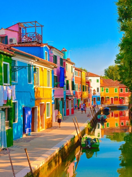 Tourists walking along the colorful houses of Burano, Venice, near a canal.