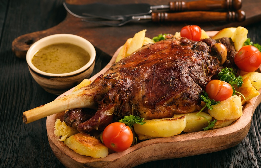 Slow baked lamb leg with potatoes and sauce on a wooden platter.