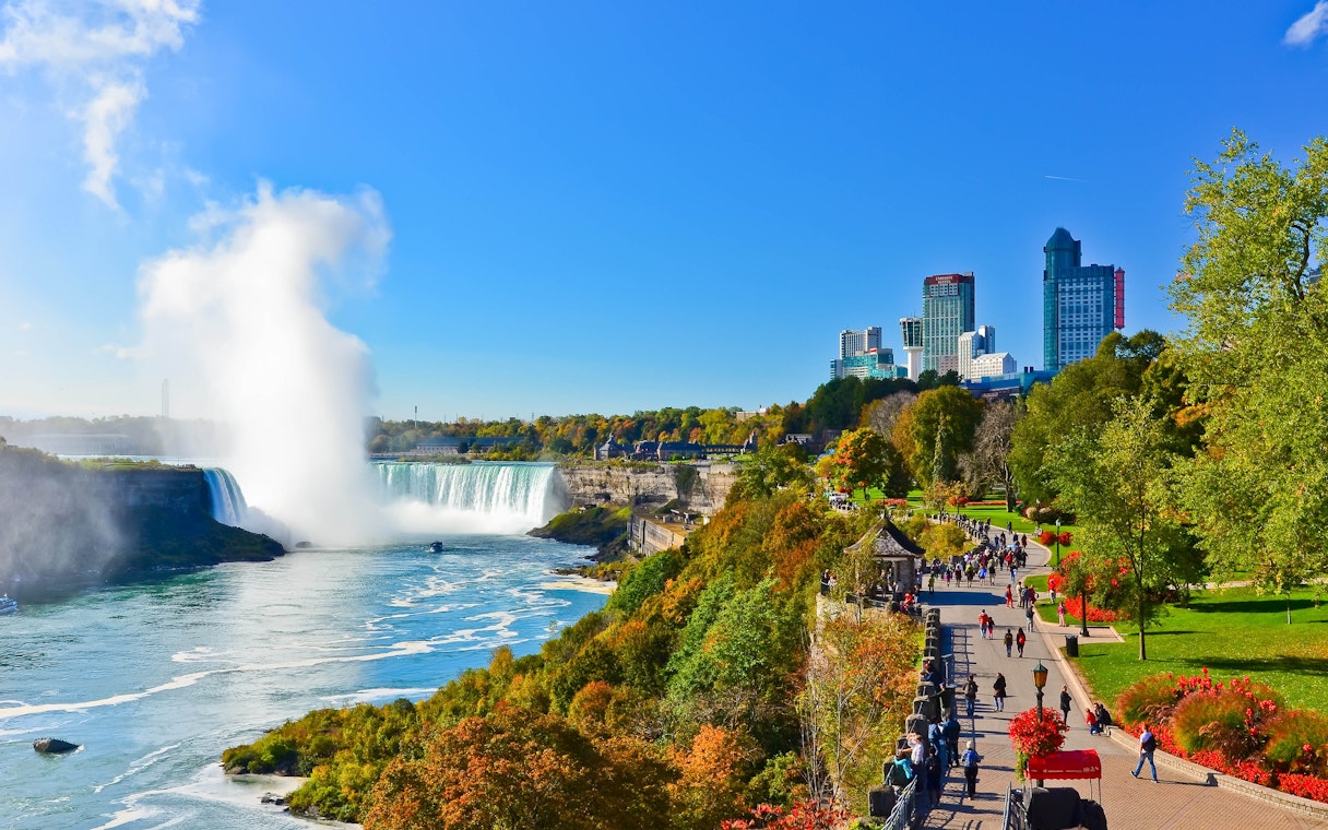Niagara Falls with city skyline and tourists walking along the park path.