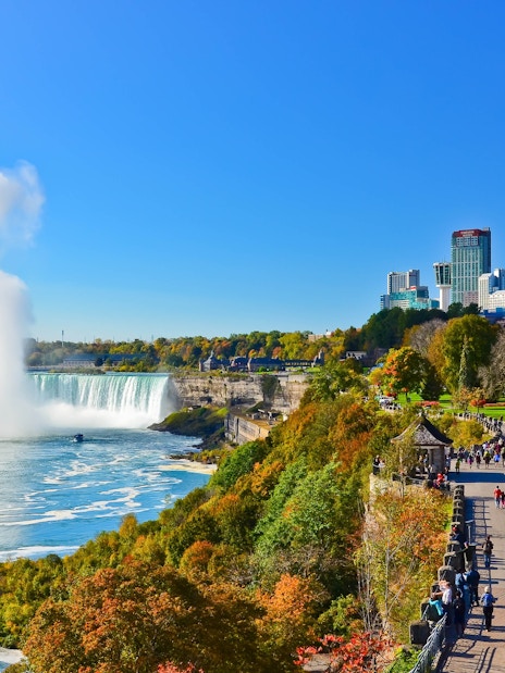 Niagara Falls with city skyline and tourists walking along the park path.