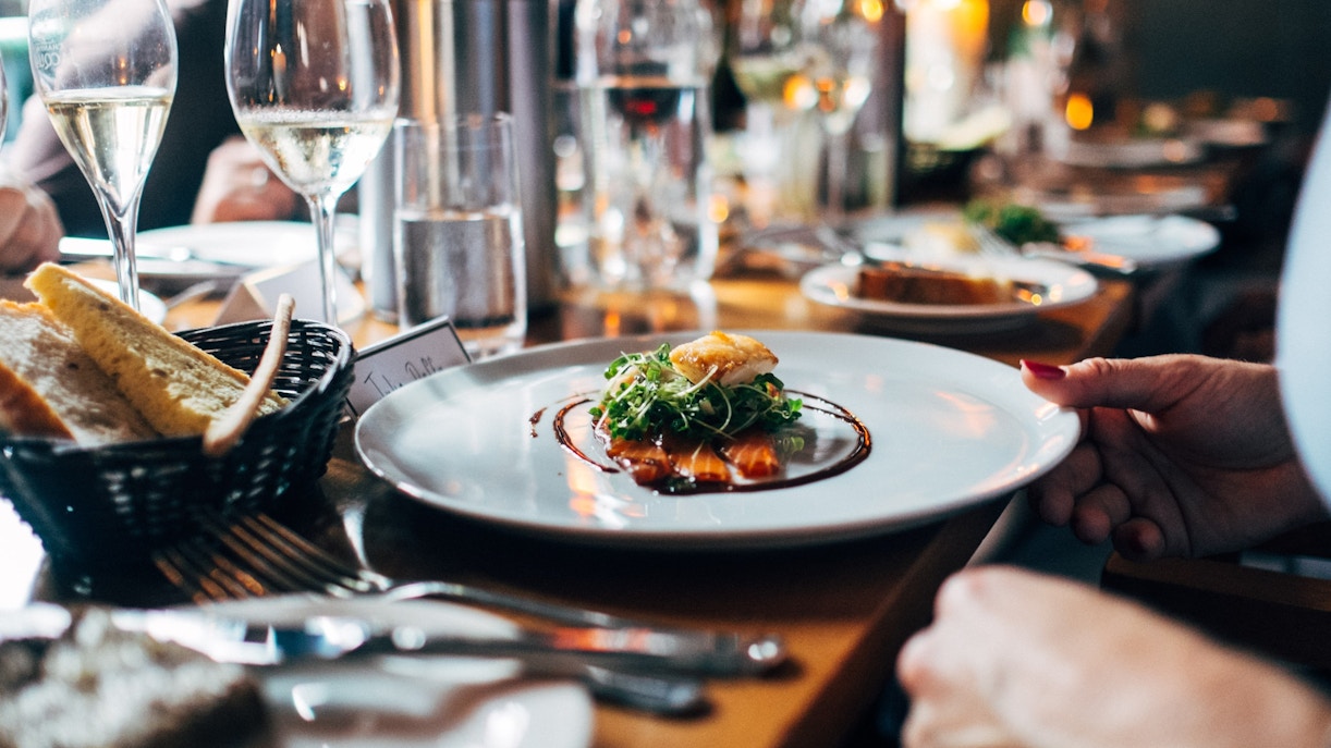Fine dining dish with greens and sauce, surrounded by wine glasses and bread basket.