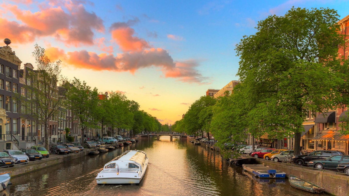 Canal cruise boat on Amsterdam canal at sunset with cityscape and trees.