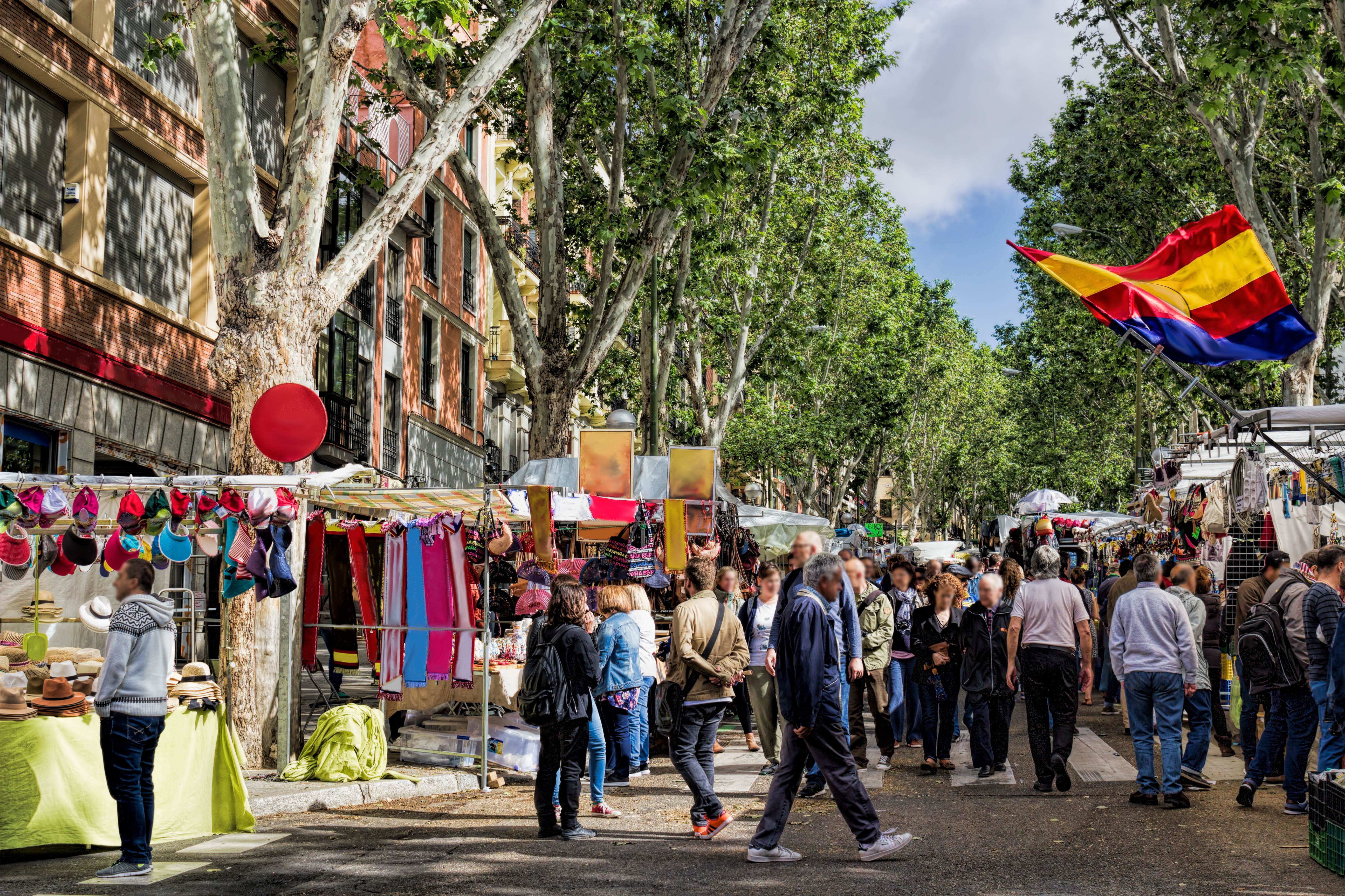 Crowded El Rastro market in Madrid with stalls selling colorful textiles and hats.