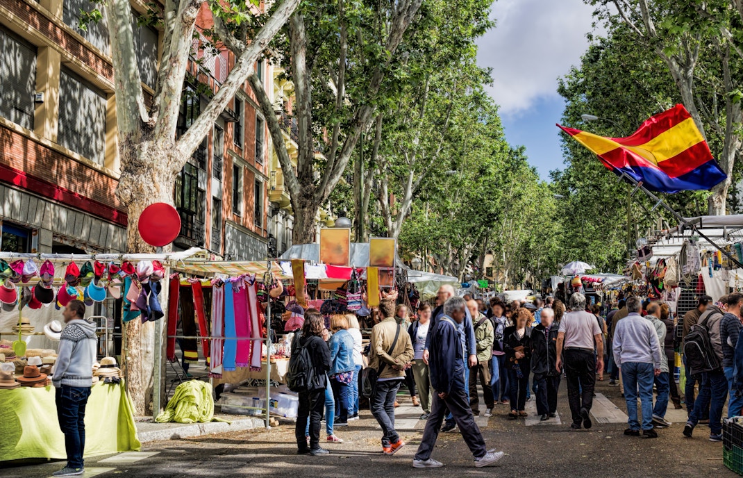 Crowded El Rastro market in Madrid with stalls selling colorful textiles and hats.