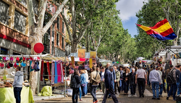 Crowded El Rastro market in Madrid with stalls selling colorful textiles and hats.