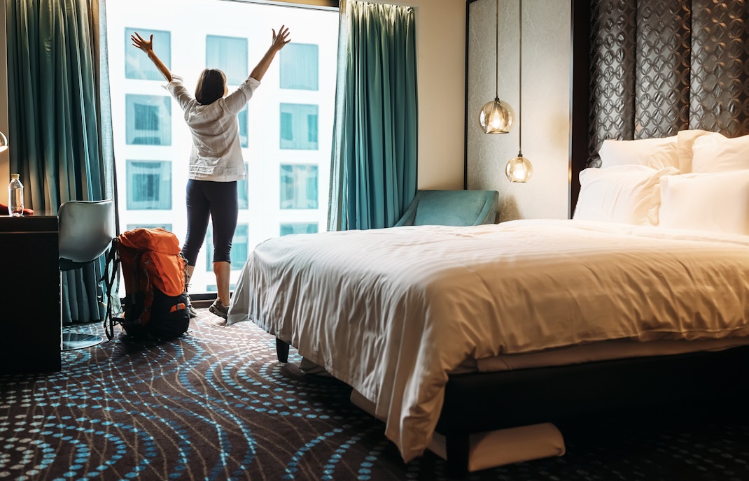 Person enjoying city view from hotel room window with backpack nearby.