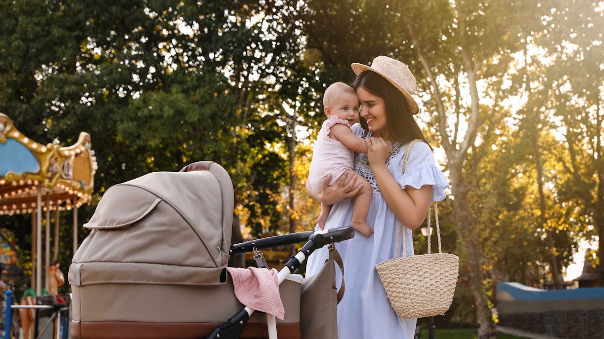 Mother holding baby next to stroller in park with carousel in background.