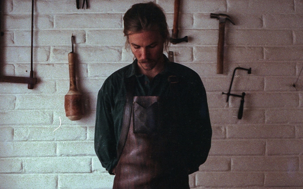 Craftsman wearing a leather apron stands in front of a brick wall with hanging tools.