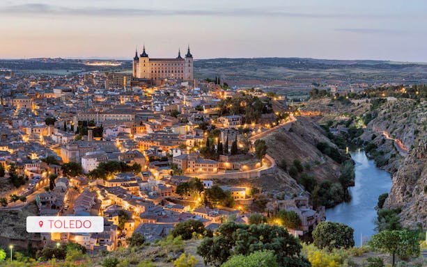 Toledo cityscape at dusk with illuminated streets and the Alcázar overlooking the Tagus River.
