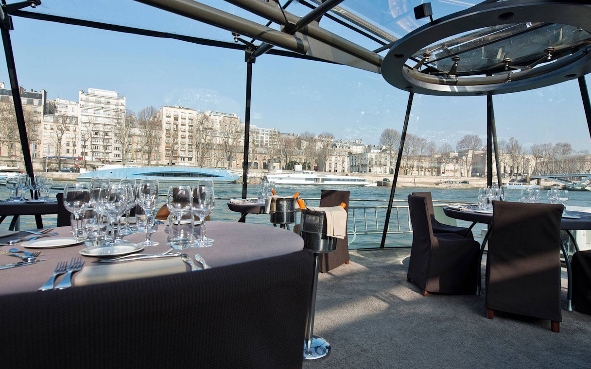 Seine River cruise dining setup with wine glasses and Parisian buildings in view.