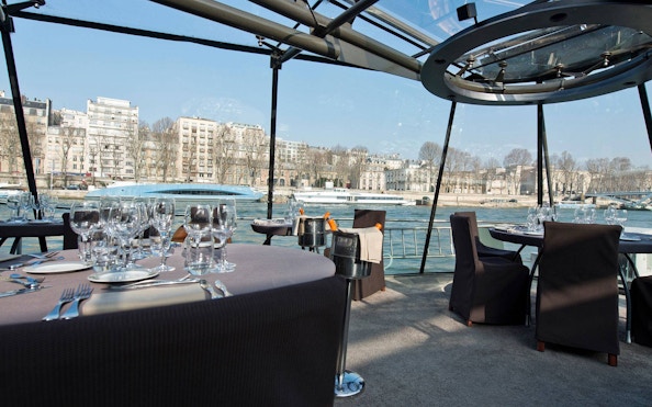 Seine River cruise dining setup with wine glasses and Parisian buildings in view.
