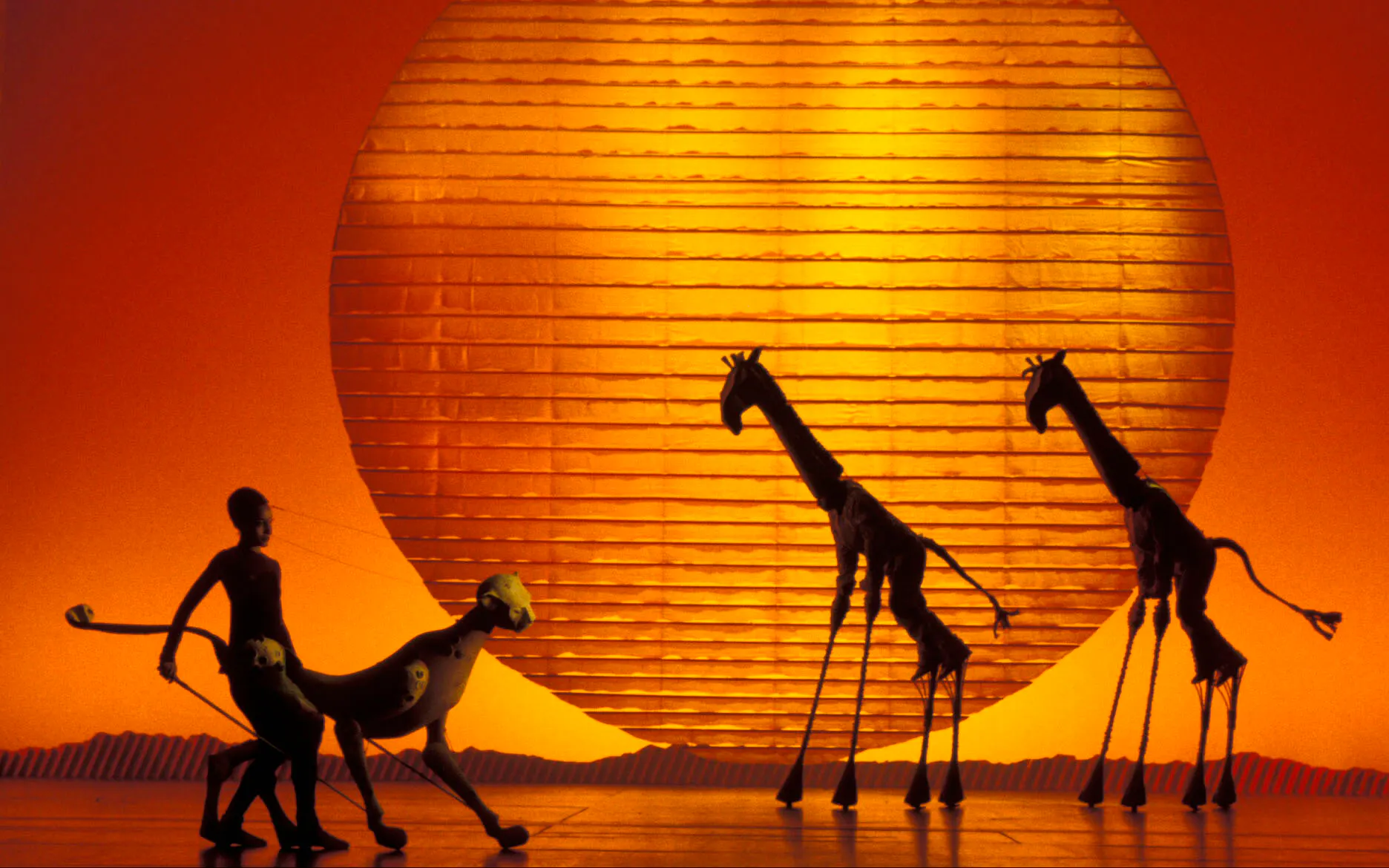 Stage performance of The Lion King with animal puppets against a sunset backdrop.