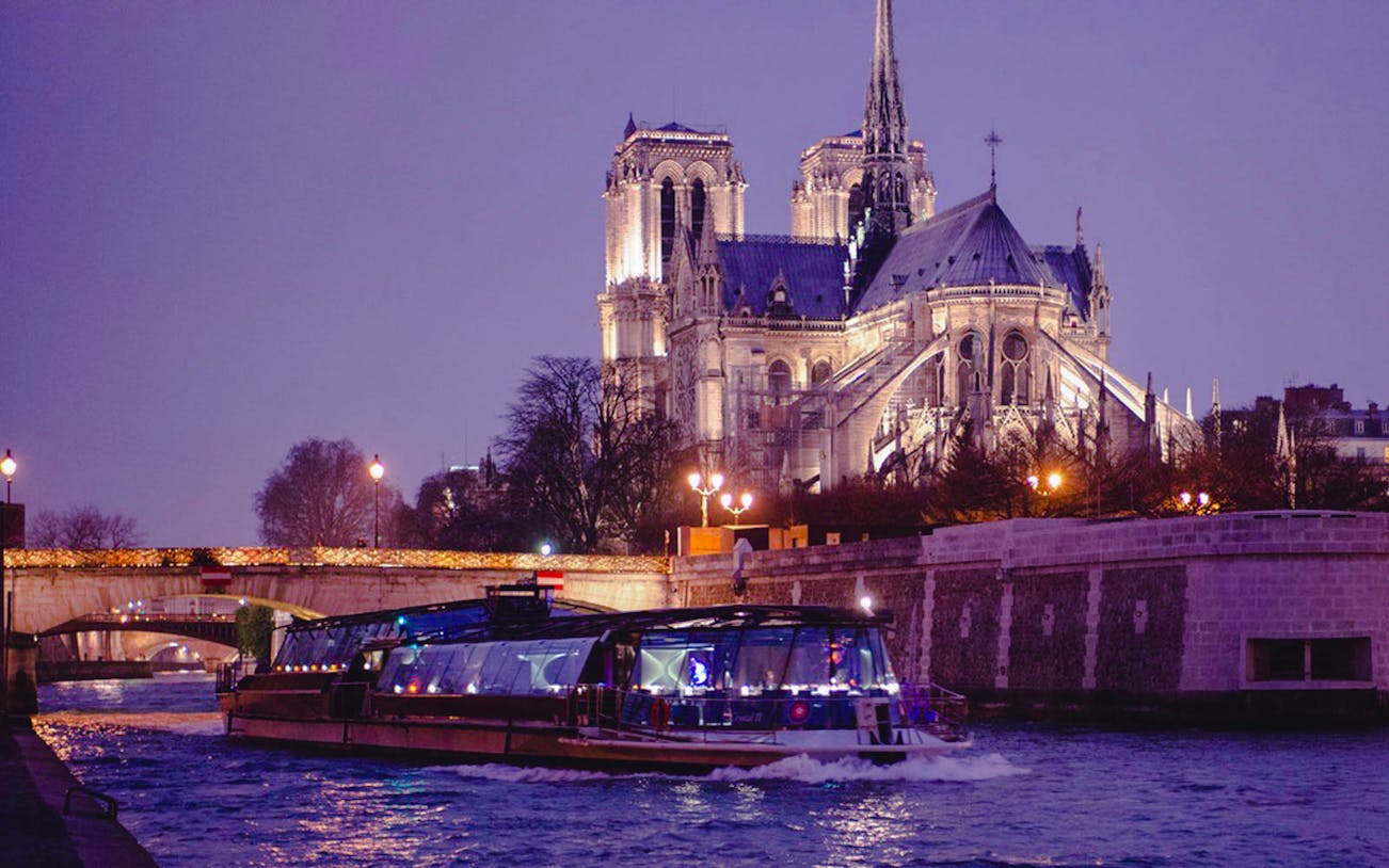Bateaux Parisiens cruise on Seine River with Notre-Dame Cathedral illuminated at night.
