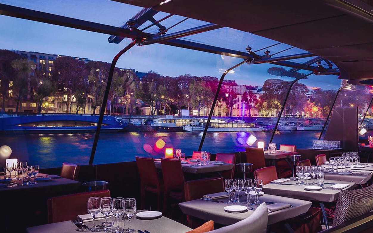 Bateaux Parisiens dinner cruise tables set with glassware, overlooking the Seine River at dusk.