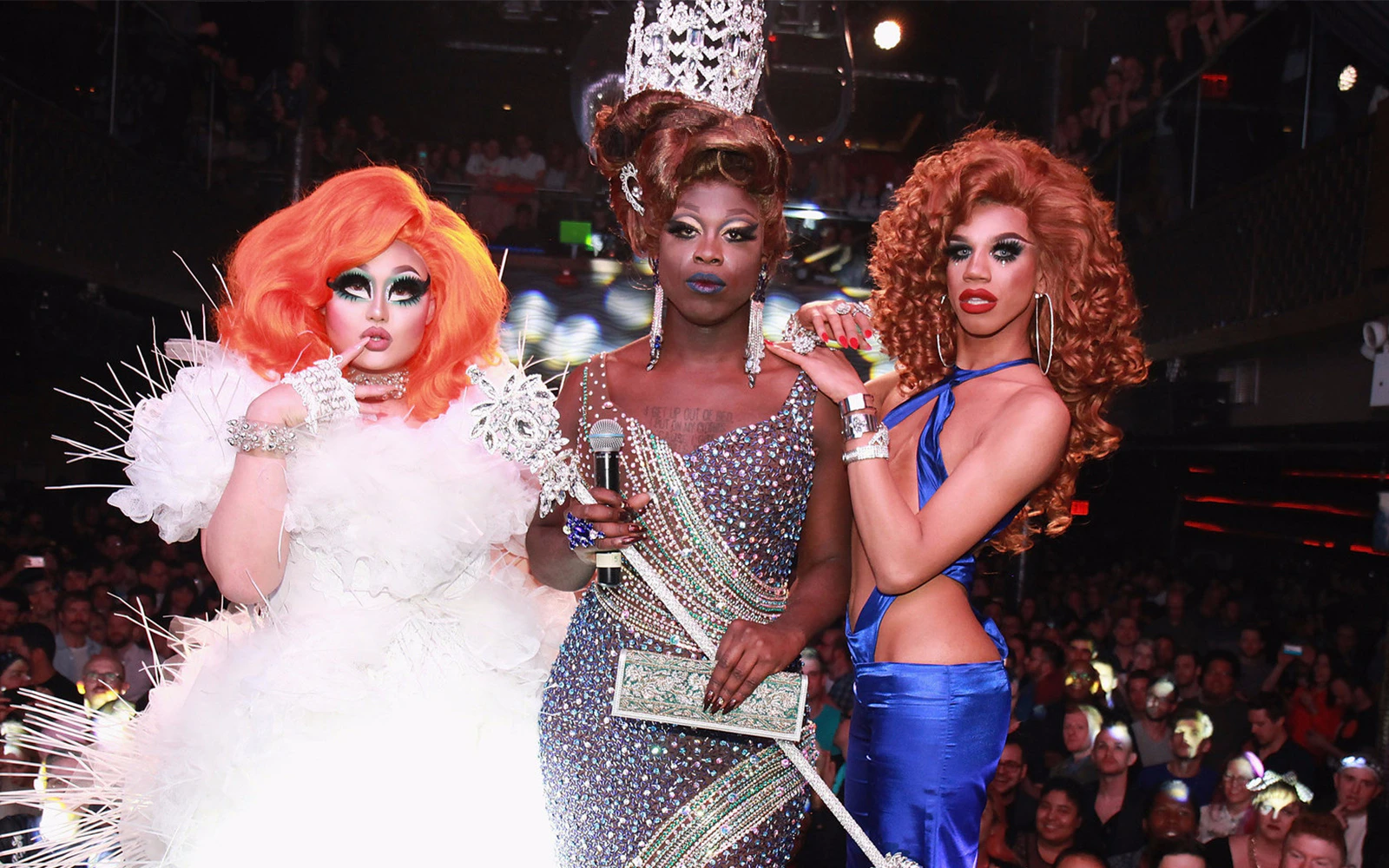 Drag queens in vibrant costumes on stage at a fashion show, holding a microphone and posing.