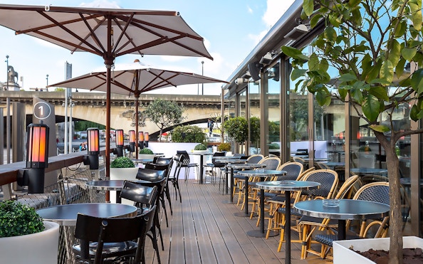 Outdoor seating at Le Bistro Parisien, Paris, with view of Seine River bridge.