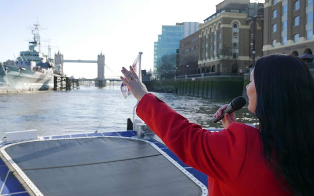 Tour guide on Thames cruise with view of Tower Bridge and HMS Belfast in London.