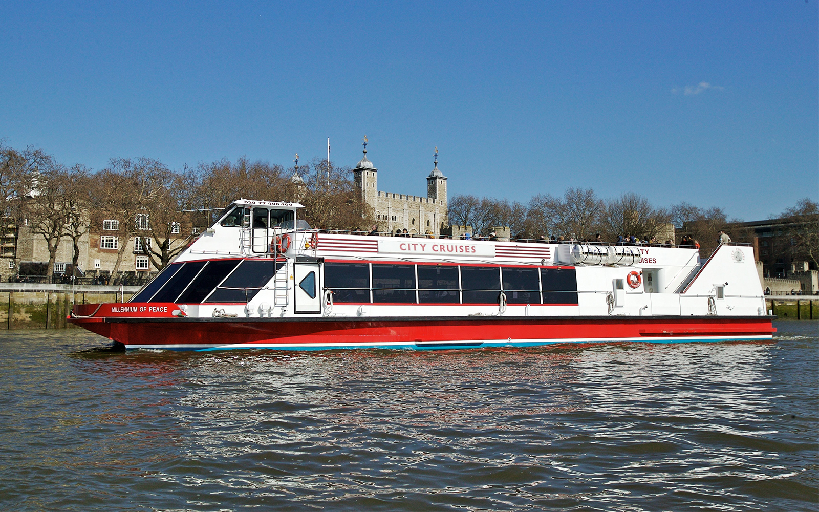 Red and white City Cruises boat on the Thames with Tower of London in the background.