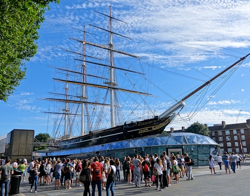 Crowd gathered near the Cutty Sark ship in Greenwich, London, under a clear blue sky.
