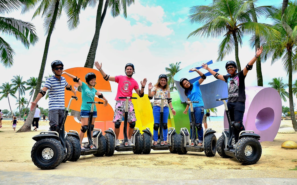 Group enjoying Segway ride on Sentosa beach with colorful letters and palm trees.