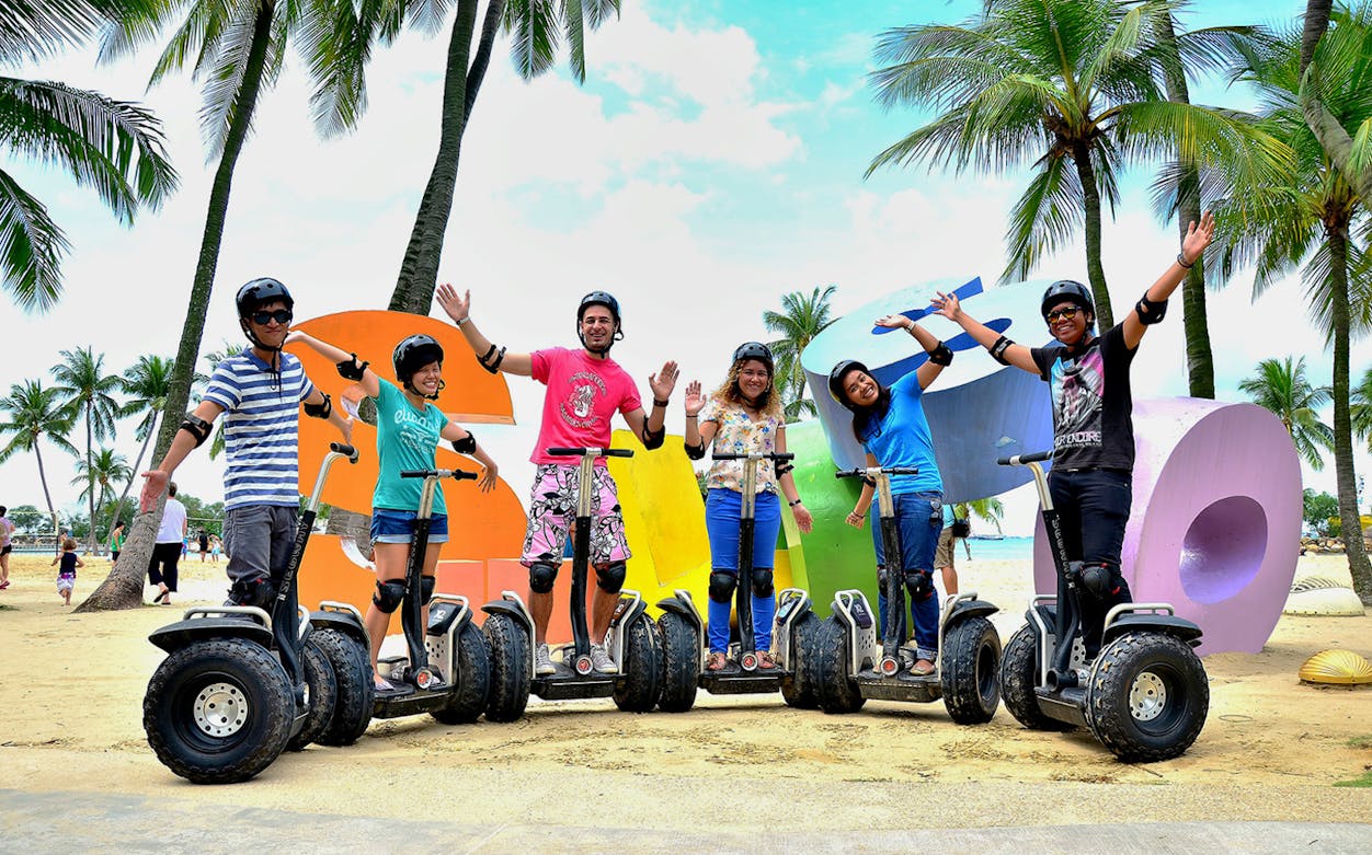 Group enjoying Segway ride on Sentosa beach with colorful letters and palm trees.