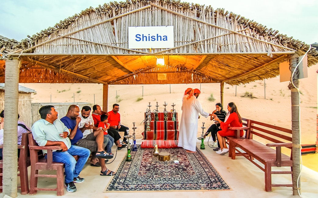Desert safari guests enjoying shisha under a thatched roof in a sandy setting.