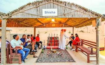 Desert safari guests enjoying shisha under a thatched roof in a sandy setting.