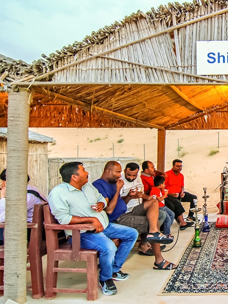 Desert safari guests enjoying shisha under a thatched roof in a sandy setting.