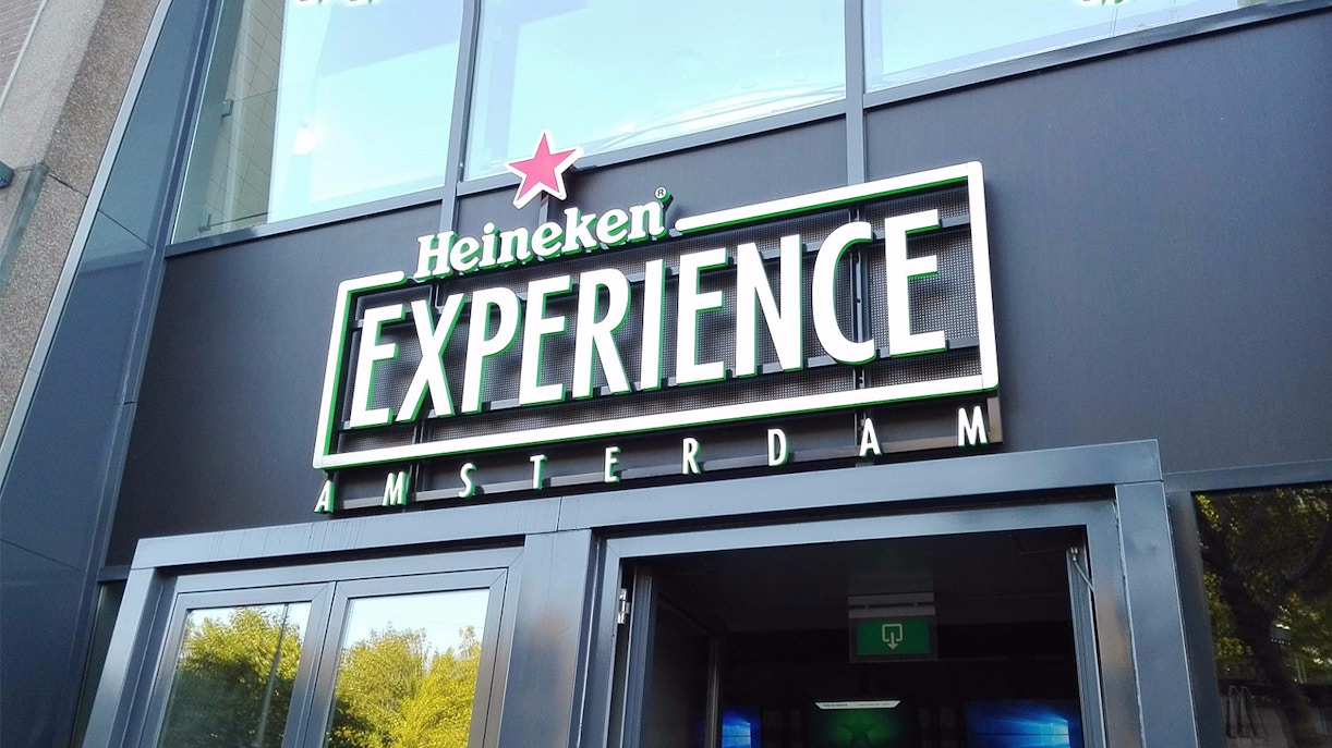 Amsterdam canal boat with tourists, Heineken Experience building in background.