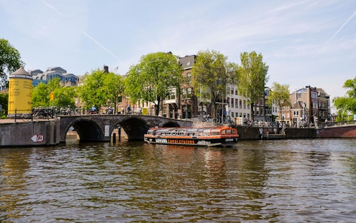 Semi-open boat cruising on Amsterdam canal near historic bridge and buildings.