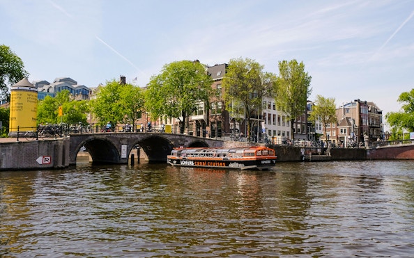 Semi-open boat cruising on Amsterdam canal near historic bridge and buildings.
