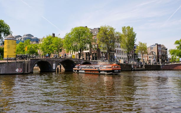 Semi-open boat cruising on Amsterdam canal near historic bridge and buildings.