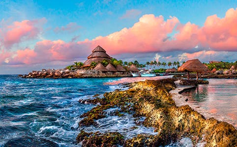 Xcaret Eco-Waterpark shoreline with traditional palapa huts and vibrant sunset sky.