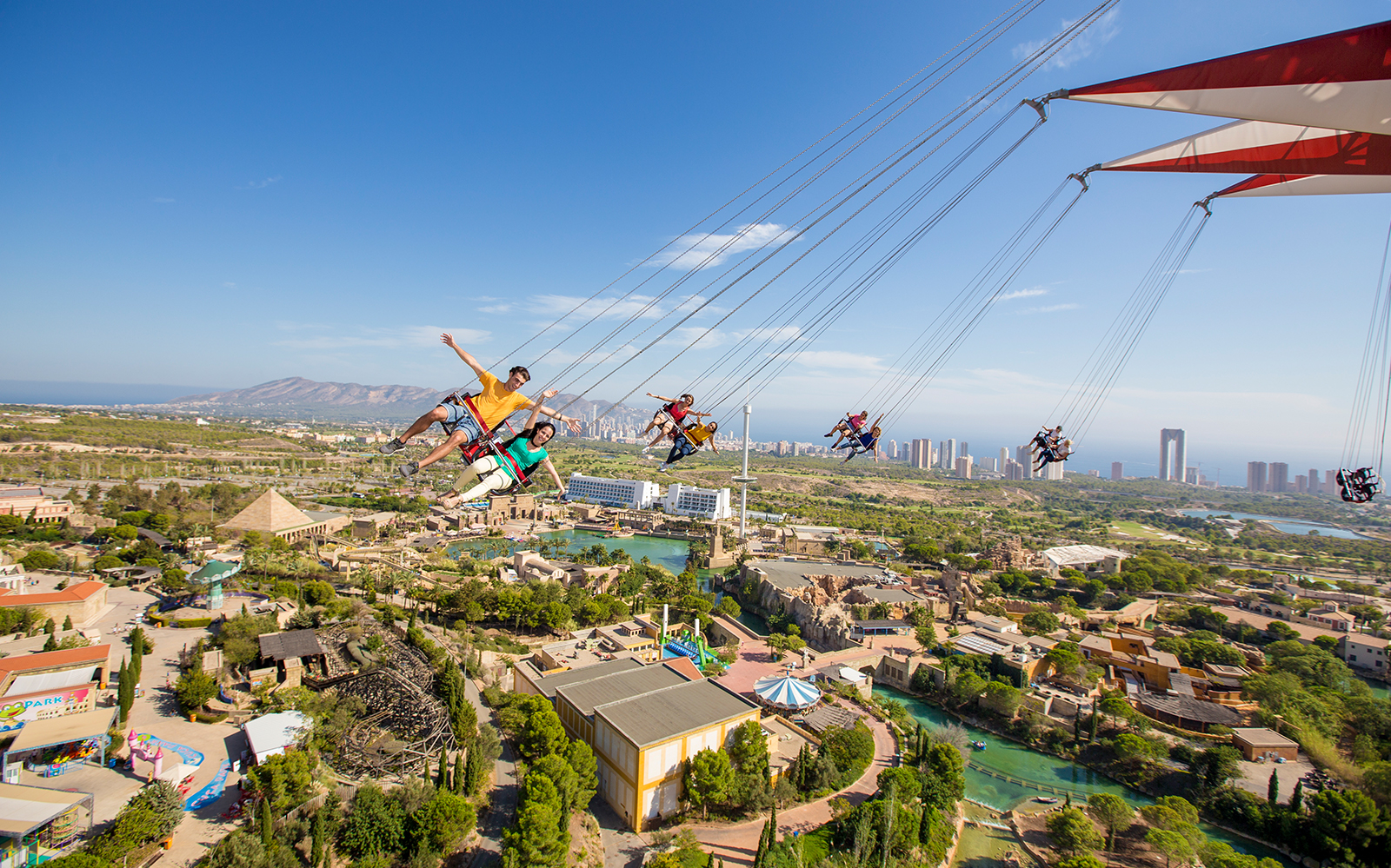 Visitors enjoying a swing ride at Terra Mitica theme park, Benidorm, with city skyline in the background.