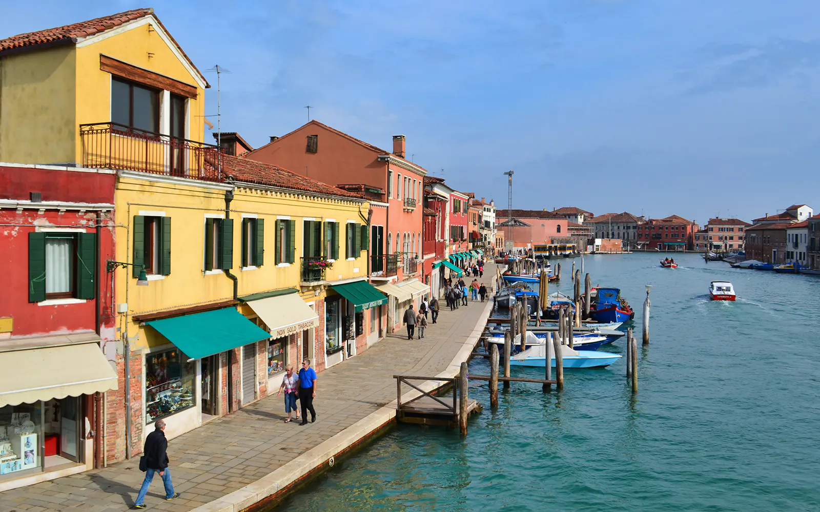 Colorful buildings along a canal in Murano, Italy, with boats and pedestrians.