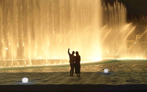 Couple watching Dubai Fountain show from boardwalk near Burj Khalifa at night.